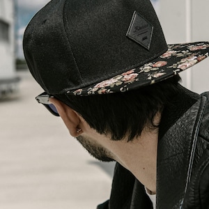 May include: Black snapback hat with a floral print brim. The hat features a small, black leather patch with a logo. The hat is made of a textured fabric and has a flat brim. The background is blurred, with a hint of blue sky.