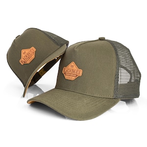 May include: Two olive green trucker hats with a mesh back and a curved brim. Each hat features a brown leather patch with the text "BLACKSKIES PREMIUM SUPPLIER MMIX". The hats are displayed on a white background.