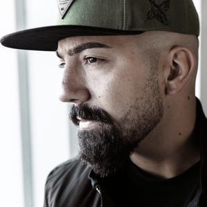 May include: Olive green and black baseball cap with a flat brim. The cap features a diamond-shaped patch with text and a bird graphic on the side. The cap is worn by a person with a beard.