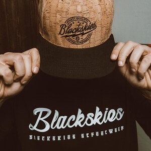 Op de afbeelding: Een bruine en zwarte baseballcap met de tekst "Blackskies" in witte letters. De cap heeft een kurkachtige textuur. Een zwarte sweatshirt met het woord "Blackskies" in witte letters is ook zichtbaar.