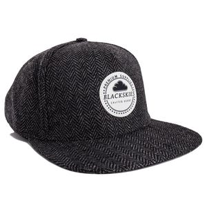 Op de afbeelding: Een zwarte en grijze baseballpet met visgraatpatroon en een platte klep. De pet heeft een cirkelvormige patch met de tekst "PREMIUM SUPPLIES BLACKSKIES CRAFTED GOODS" in wit op een zwarte achtergrond.