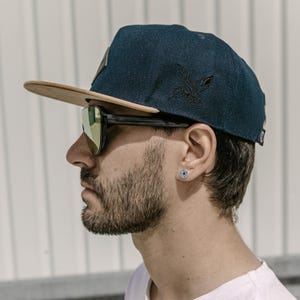 Könnte beinhalten: Eine dunkelblaue Baseballkappe aus Denim mit flachem beigefarbenem Schirm und einem schwarzen, gestickten Vogelmotiv an der Seite. Die Kappe hat einen kleinen Aufnäher auf der Vorderseite und ein kreisförmiges Logo hinten. Getragen mit einer schwarzen Sonnenbrille.