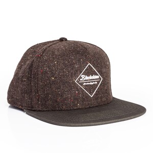 Op de afbeelding: Bruine tweed baseballcap met een platte klep. De pet heeft een wit ruitvormig logo met het woord "Blackshore". De tweed stof heeft rode, gele en witte vlekken.