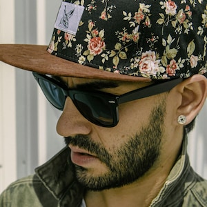 Puede incluir: Gorra de béisbol negra con un estampado floral de rosas rojas y rosas y hojas verdes. La gorra tiene una visera de ante marrón y un parche cuadrado blanco con un gráfico negro. Gafas de sol negras.