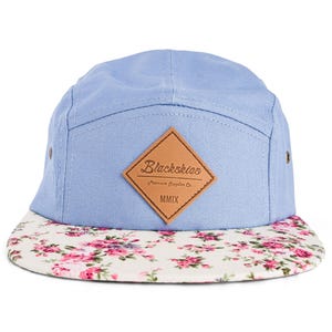 Op de afbeelding: Een lichtblauwe vijf-panel cap met een bloemenprint klep. De cap heeft een bruin leren patch met de tekst "Blackskies" en "MMIX". De klep heeft een witte achtergrond met roze en rode rozen.