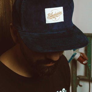 Op de afbeelding: Marineblauwe corduroy baseballcap met een vierkante patch met de tekst "Blackskies" in gouden letters. De cap heeft een gebogen klep en een gestructureerde kroon. Een modieus accessoire.