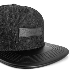 Könnte beinhalten: Schwarze Denim-Baseballkappe mit schwarzem Lederschild. Die Kappe hat einen rechteckigen Lederaufnäher mit dem geprägten Wort "Blackskies". Ein stilvolles Accessoire.