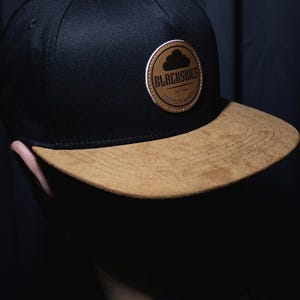 Könnte beinhalten: Schwarze Baseballkappe mit braunem Wildleder-Schirm. Die Kappe hat einen runden Aufnäher mit dem Text "BLACKSKIES" und einer Wolkengrafik. Ein Premium-Accessoire.