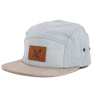 Puede incluir: Gorra de cinco paneles de mezclilla azul claro con visera de ante beige. Un parche de cuero marrón en la parte delantera presenta una ilustración negra. La gorra tiene dos pequeños ojales metálicos en el lateral.