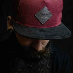 May include: A maroon baseball cap with a flat black brim and a diamond-shaped black patch on the front. The patch features embossed text that reads Blackskies, Premium Quality Co, and MMXII. The cap has a structured crown and a button on top.