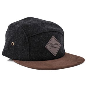 Può includere: Un cappellino a cinque pannelli nero e marrone con visiera piatta. Il cappellino presenta una toppa in pelle a forma di diamante con la scritta "Blueberries" e un piccolo occhiello metallico. La visiera è marrone.