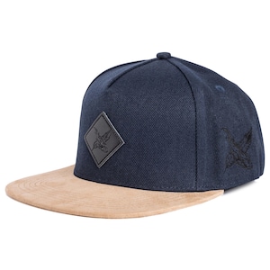 May include: A navy blue flat-peak cap with a tan suede peak. The cap has a black diamond-shaped patch with a bird design on the front and a bird graphic on the side.