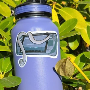 May include: A purple stainless steel water bottle with a black lid and a carrying handle. The bottle has a sticker of a video game console with a snake wrapped around it.