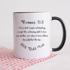 May include: White ceramic mug with a black handle and rim, featuring the text "Romans 13:8" and a Bible verse. The mug also has the words "100% Debt Free" in a script font. The mug is on a red and white checkered surface.
