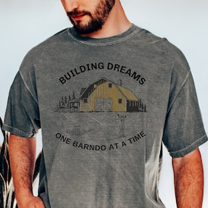 May include: A gray t-shirt with a distressed look features a graphic of a yellow barn with the text "Building Dreams One Barndo at a Time".