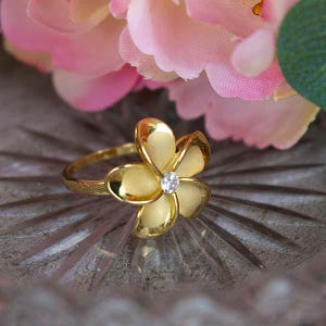 May include: A gold-colored ring featuring a flower design with five petals. The center of the flower has a small, clear gemstone. The ring is set against a blurred background of pink flowers.