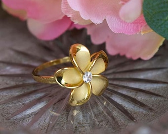 May include: A gold-colored ring featuring a flower design with five petals. The center of the flower has a small, clear gemstone. The ring is set against a blurred background of pink flowers.