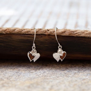 Puede incluir: Un par de pendientes de plata con dos colgantes en forma de corazón. El corazón más pequeño está hecho de madera y es de color marrón. El corazón más grande es de plata.