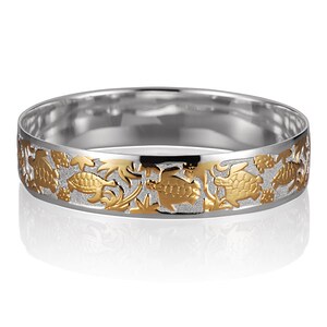 Puede incluir: Un brazalete de plata con un diseño dorado de tortugas marinas y plantas. El brazalete está pulido y tiene un acabado brillante.