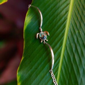 Può includere: Braccialetto d'argento con un ciondolo a forma di tartaruga in madreperla verde e blu. Il braccialetto è su una foglia verde.