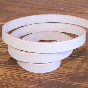 May include: A long, continuous strip of natural, undyed vegetable-tanned leather coiled into a spiral shape. The leather strap features a smooth, light beige surface with visible grain texture and raw, unfinished edges along the entire length.