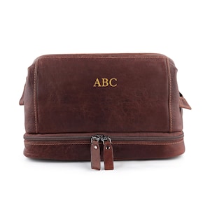 May include: A brown leather dopp kit with a dual zipper design. The bag features the gold-colored initials "ABC" on the front. The bag has a rectangular shape with a textured surface and a handle on each side.