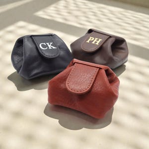 Könnte beinhalten: Drei kleine, weiche Beutel in Schwarz, Dunkelbraun und Terrakotta. Jeder Beutel verfügt über eine Umschlagklappe mit Druckknopfverschluss. Der schwarze Beutel ist mit weißen Buchstaben CK geprägt, der braune mit goldenen Buchstaben PH, der terrakottafarbene ist schlicht.