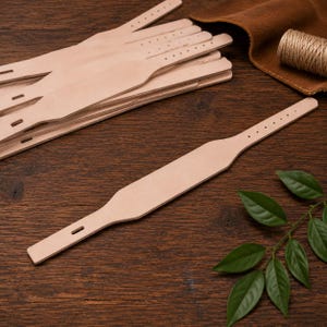May include: Several unfinished, pale tan leather straps are visible, each with a line of small holes. One strap lies in the foreground. Green leaves are in the bottom right corner. The straps are on a dark brown wooden surface.