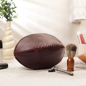 May include: A brown leather toiletry bag shaped like a football, with stitched detailing. A shaving brush, razor, and other grooming items are arranged nearby. The bag is approximately 12 inches long.