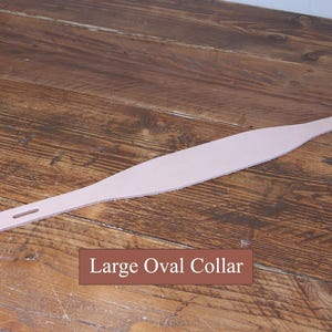 Peut inclure: Un collier en cuir rose clair avec une partie centrale large et ovale qui se r&eacute;tr&eacute;cit vers les extr&eacute;mit&eacute;s. Une extr&eacute;mit&eacute; pr&eacute;sente une fente rectangulaire, tandis que l'autre poss&egrave;de une s&eacute;rie de petits trous circulaires. Le texte Large Oval Collar est visible.