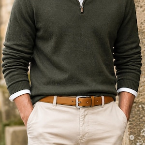 May include: A dark olive green quarter-zip sweater worn over a white shirt, paired with beige trousers and a tan leather belt featuring a silver-toned metal buckle. The sweater has long sleeves with ribbed cuffs, and the belt is threaded through loops.