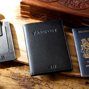 May include: A black leather passport holder with the word "PASSPORT" embossed on the front. The initials "SB" are embossed at the bottom. A gray cardholder and a British passport are also visible.