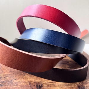May include: Close-up of four leather strips in shades of red, navy blue, black, and brown. The strips are curled and layered, showcasing the texture and color variations of the leather. The image highlights the different colors and the material's flexibility.
