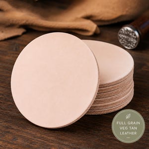 Pode incluir: Uma pilha de bases redondas de couro bege claro. As bases são feitas de couro de flor integral, curtido vegetal. Uma ferramenta de carimbo é visível no fundo. As bases estão em uma superfície de madeira escura.
