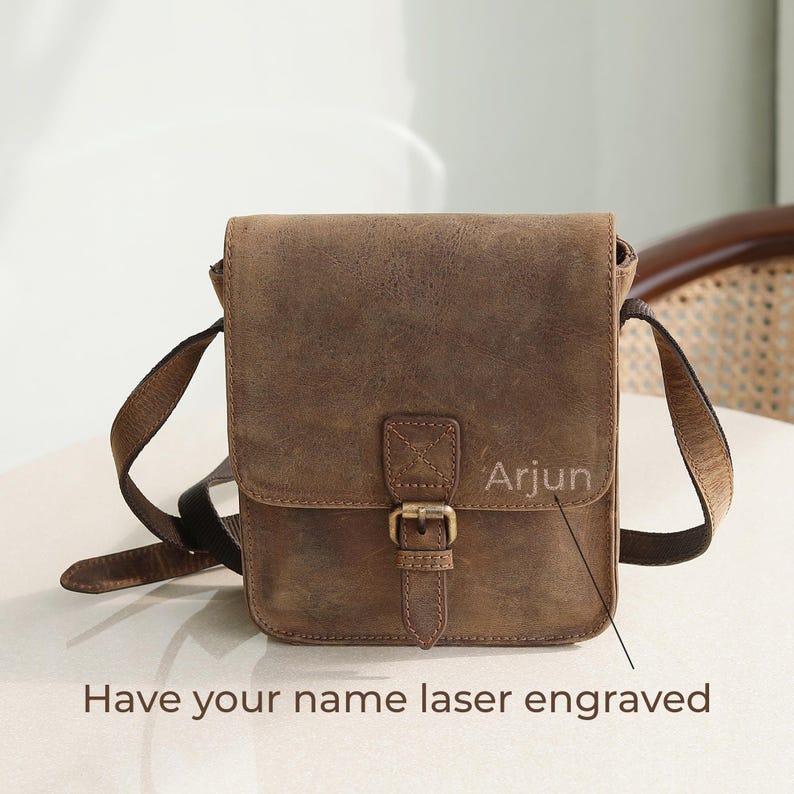 Op de afbeelding: Een bruine leren schoudertas met een voorflap, een enkele gespsluiting en een verstelbare schouderriem. De tas heeft de naam Arjun in de voorflap gegraveerd, met de tekst Have your name laser engraved eronder weergegeven.