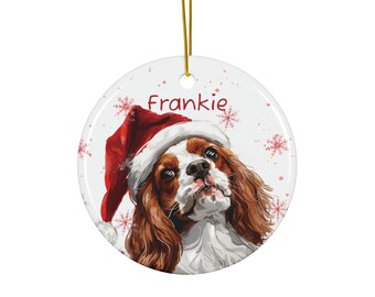 May include: A round, white Christmas ornament featuring a Cavalier King Charles Spaniel wearing a Santa hat. The dog's name, "Frankie," is written in red script above the dog's head. Red snowflakes are scattered around the edges.