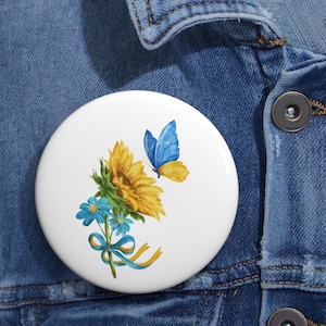 Op de afbeelding: Een witte knop met een zonnebloem, blauwe bloemen en een blauwe vlinder met gele vleugels. De zonnebloem en de vlinder zijn met een blauw en geel lint aan elkaar gebonden.