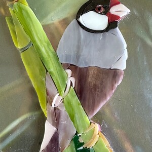 Op de afbeelding: Een moza&iuml;ekkunstwerk van een kleine vogel die op een groene bamboestok zit. De vogel is gemaakt van glazen tegels in tinten grijs, bruin, wit en rood. De achtergrond is een wazig groen en bruin.