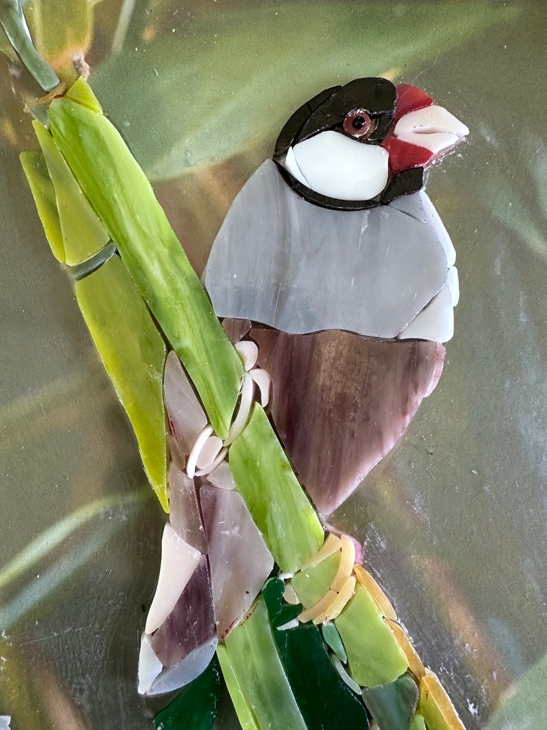 Op de afbeelding: Een moza&iuml;ekkunstwerk van een kleine vogel die op een groene bamboestok zit. De vogel is gemaakt van glazen tegels in tinten grijs, bruin, wit en rood. De achtergrond is een wazig groen en bruin.