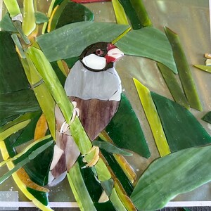 Op de afbeelding: Een moza&iuml;ekkunstwerk van een kleine grijze en bruine vogel die op een groene bamboestok zit. De vogel heeft een rode snavel en een witte vlek op zijn borst. De achtergrond is een groene en gele moza&iuml;ek van bladeren.