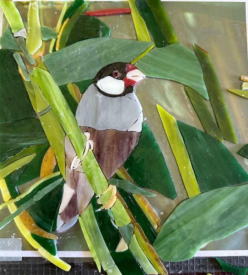 Op de afbeelding: Een moza&iuml;ekkunstwerk van een kleine grijze en bruine vogel die op een groene bamboestok zit. De vogel heeft een rode snavel en een witte vlek op zijn borst. De achtergrond is een groene en gele moza&iuml;ek van bladeren.