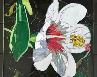 Op de afbeelding: Een witte bloem met rode en groene accenten is gemaakt van mozaïektegels. De bloem is geplaatst tegen een zwarte achtergrond.