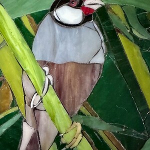 Op de afbeelding: Een glas-in-lood moza&iuml;ek van een vogel die op een groene tak zit. De vogel heeft een grijs lichaam, een zwarte kop en een rode snavel. De achtergrond is een groene en gele moza&iuml;ek.