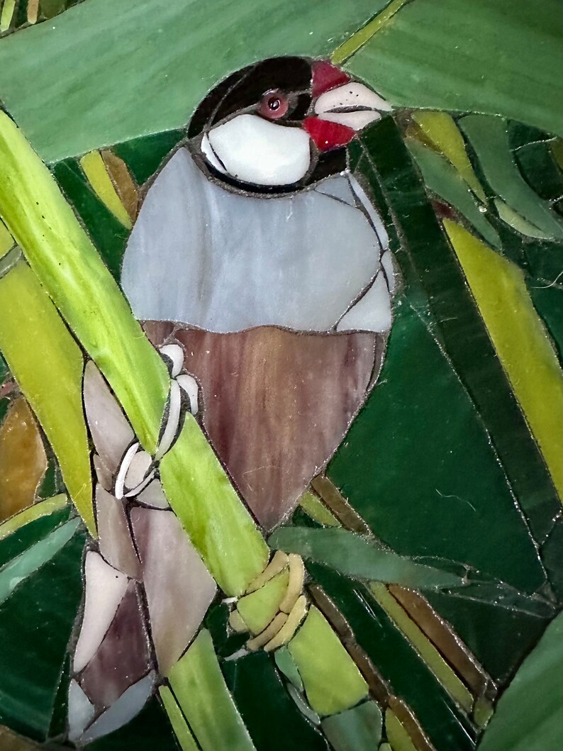Op de afbeelding: Een glas-in-lood moza&iuml;ek van een vogel die op een groene tak zit. De vogel heeft een grijs lichaam, een zwarte kop en een rode snavel. De achtergrond is een groene en gele moza&iuml;ek.