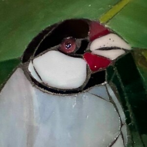 Op de afbeelding: Close-up van een vogel van gekleurd glas met een rode snavel, een zwarte kop en een witte borst. De vogel is omringd door groen en geel gekleurd glas.