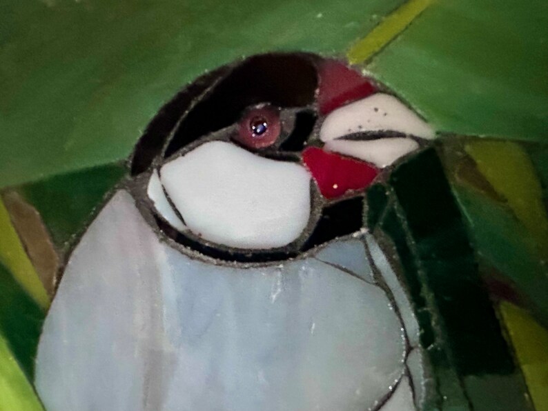 Op de afbeelding: Close-up van een vogel van gekleurd glas met een rode snavel, een zwarte kop en een witte borst. De vogel is omringd door groen en geel gekleurd glas.