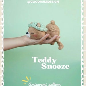 Può includere: Amigurumi di un orsacchiotto all'uncinetto in posizione di riposo, con un cappello verde chiaro. L'orso è tenuto in una mano su uno sfondo verde chiaro. Il testo recita "Teddy Snooze" e "Amigurumi pattern".