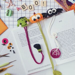 Può includere: Tre ciondoli per segnalibro di Halloween all'uncinetto. Un Frankenstein verde, una zucca arancione e un ragno nero. I segnalibro sono appesi a un libro con il testo "XVII" sulla pagina. Il testo "BOO!" è visibile sullo sfondo.