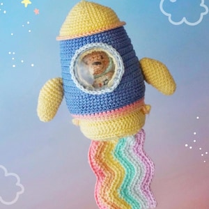 Può includere: Razzo all'uncinetto fatto a mano in blu, giallo e rosa. Il razzo ha una finestra trasparente con un orsacchiotto all'interno. Lo scarico del razzo è un arcobaleno di colori. Il design include piccole ali e una parte superiore arrotondata.