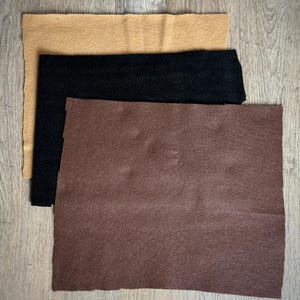May include: Three pieces of felt fabric in different colors: brown, black, and tan. The fabric is laid out in a stack on a wooden surface.